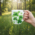 Hand holding a mug with a four leaf clover design in a forest setting.